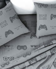The Juego de Sábanas Play by Intima Hogar features a modern, gamer-themed design with a gray base, video game controller graphics, and fun phrases like GAME OVER, PRESS START, CONTINUE?, plus pixelated hearts. Este set incluye dos fundas de almohada y sábanas perfectas para los fans de los videojuegos.
