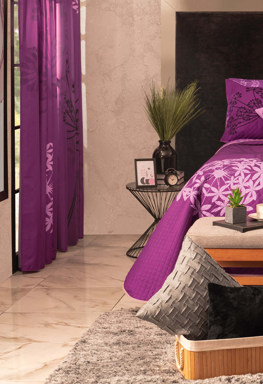 A bedroom corner features a modern black side table with decor, a bed with purple floral bedding, and Juego de Cortinas Violeta by Intima Hogar. The space includes a gray rug, a cushioned bench, and a marble floor.