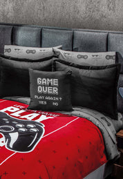 The Juego de Sábanas Play by Intima Hogar features black and red microfibra sheets with a game controller design. The center pillow says “Game Over. Play again? Yes No.” The bed has a gray headboard and a concrete-like textured wall.