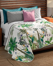 A neatly made bed features the Intima USA Juego de Edredón Tropical with green leaves and exotic flowers over a gray quilted cover, accented by teal, gray, and pink pillows. Wooden nightstands and a woven rug complete the look.