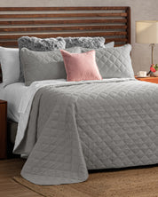 A neatly made bed features the Intima USA Juego de Edredón Tropical in light gray, complemented by matching pillows, a pink accent pillow, and two fluffy gray ones. The wooden headboard stands next to a nightstand with a lamp and teacup.