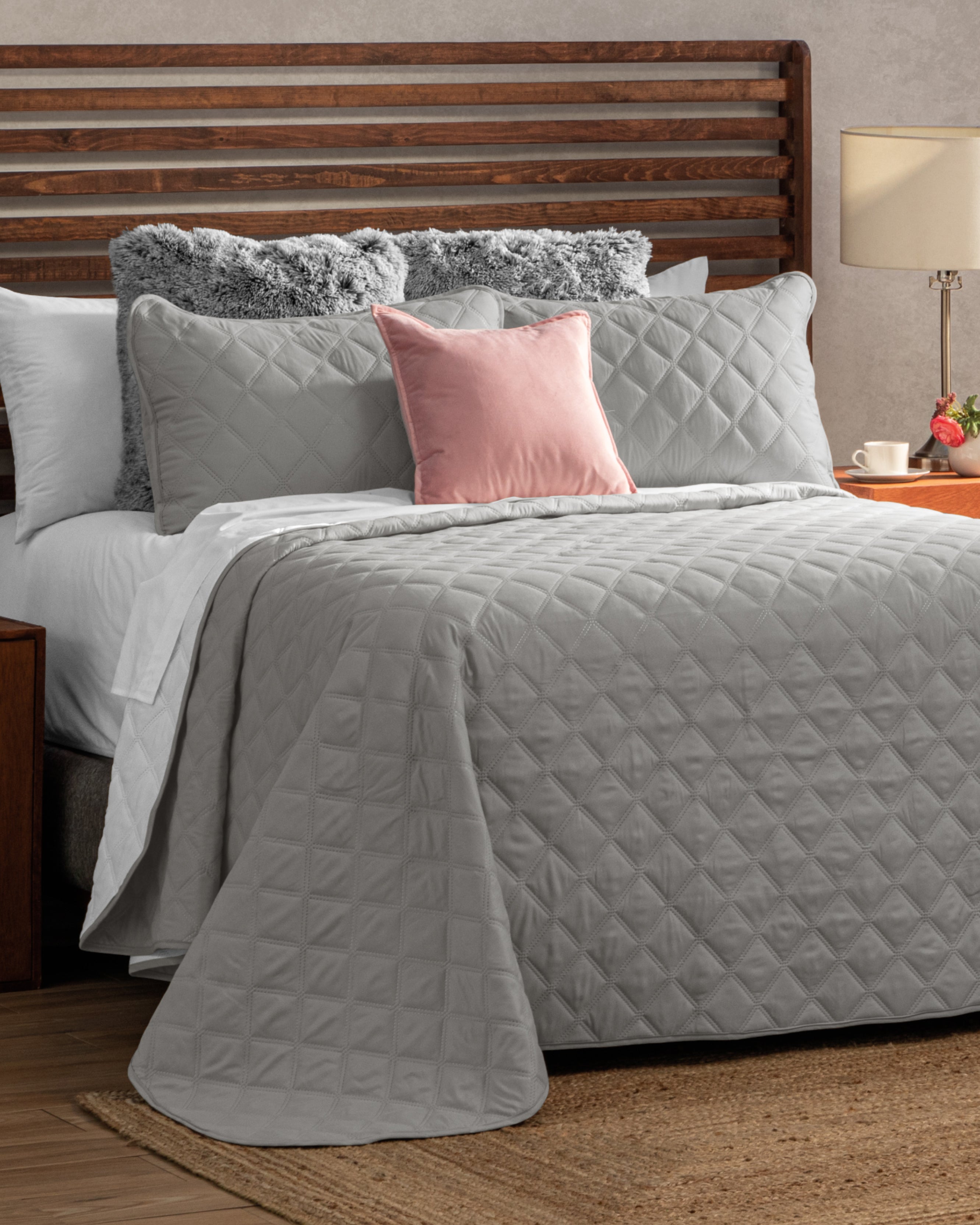 A neatly made bed features the Intima USA Juego de Edredón Tropical in light gray, complemented by matching pillows, a pink accent pillow, and two fluffy gray ones. The wooden headboard stands next to a nightstand with a lamp and teacup.