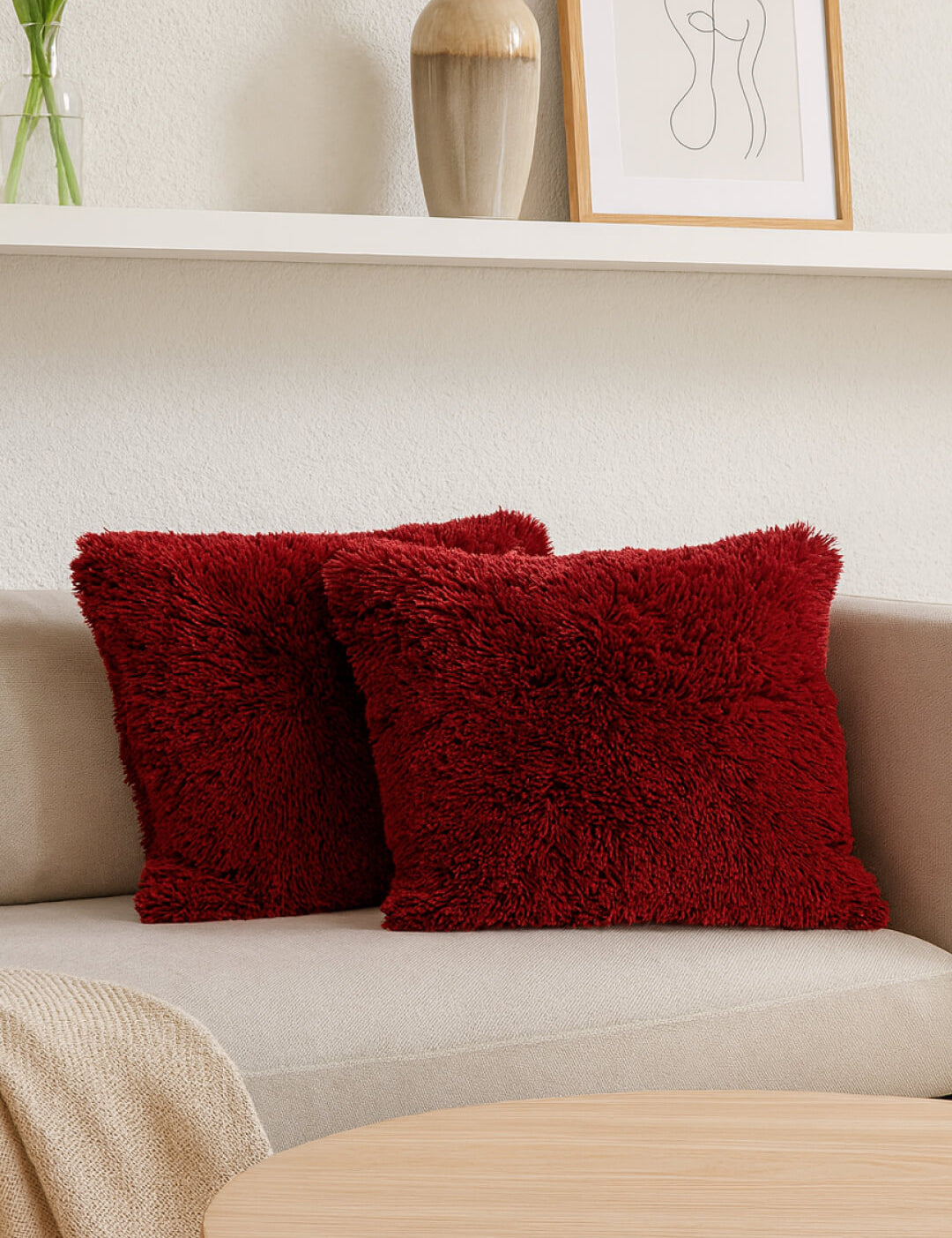 Dos cojines del Juego de Cojines de Lujo Africa de Intima Hogar en rojo reposan sobre un sofá beige claro. Una manta color tan cubre el brazo, frente a una mesa de centro; al fondo se ve un estante con jarrón, planta y arte abstracto.