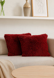 Dos cojines del Juego de Cojines de Lujo Africa de Intima Hogar en rojo reposan sobre un sofá beige claro. Una manta color tan cubre el brazo, frente a una mesa de centro; al fondo se ve un estante con jarrón, planta y arte abstracto.