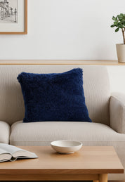 El Juego de Cojines de Lujo Denver de Intima Hogar en azul marino decora un sofá beige claro en una sala moderna, acompañado de mesa de centro de madera, libro abierto, tazón blanco, cuadro en la pared y planta en una repisa.