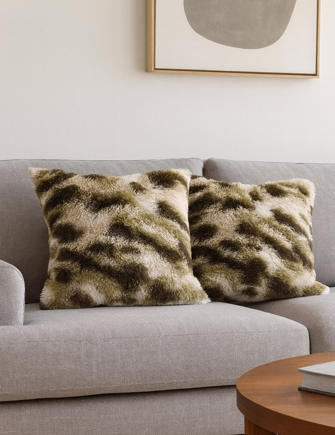 Dos cojines Juego de Cojines de Lujo Finlandia de Intima Hogar, en tonos verde y beige, decoran un sofá gris claro en una sala moderna con mesa de madera y arte abstracto.