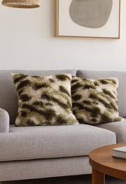 Dos cojines Juego de Cojines de Lujo Finlandia de Intima Hogar, en tonos verde y beige, decoran un sofá gris claro en una sala moderna con mesa de madera y arte abstracto.