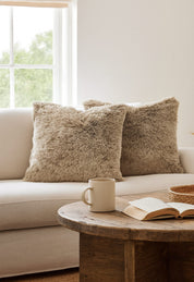 Dos cojines del Juego de Cojines de Lujo Glacial de Intima Hogar en tono beige decoran un sofá claro junto a la ventana, mientras una mesa de centro sostiene un libro abierto, una taza beige y una bandeja tejida para un ambiente acogedor.