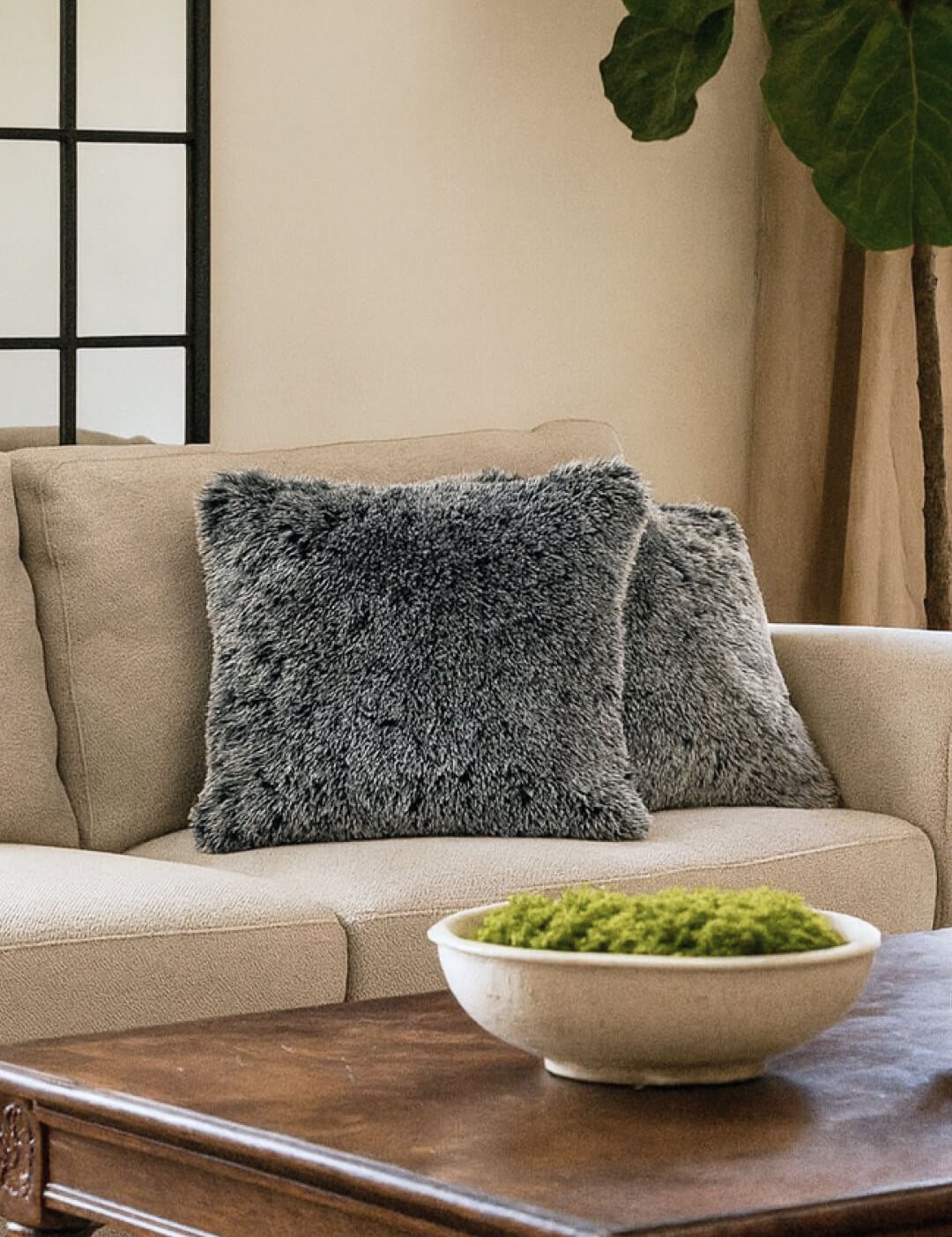A beige sofa with two Juego de Cojines de Lujo Humo by Intima Hogar in gray sits beside a large plant. In front, a wooden coffee table holds a white bowl of green moss. Behind the sofa, a window with black grid panes is visible.