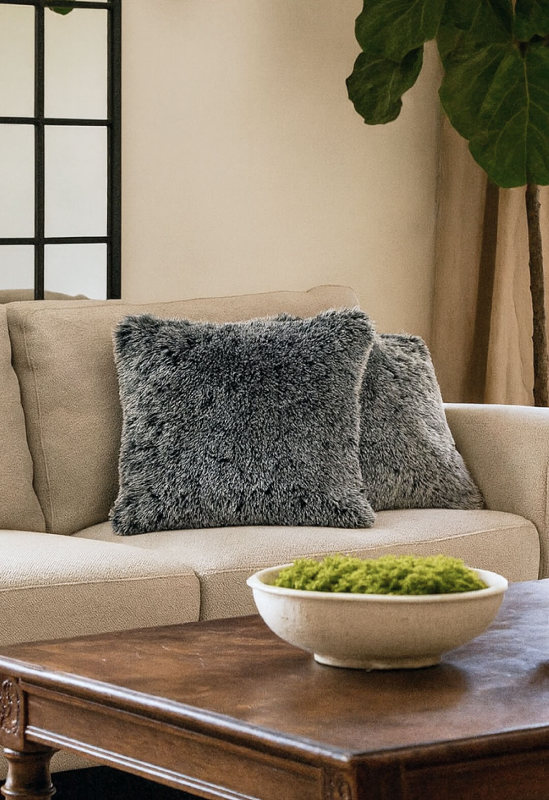 A beige sofa with two Juego de Cojines de Lujo Humo by Intima Hogar in gray sits beside a large plant. In front, a wooden coffee table holds a white bowl of green moss. Behind the sofa, a window with black grid panes is visible.