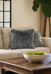 A beige sofa with two Juego de Cojines de Lujo Humo by Intima Hogar in gray sits beside a large plant. In front, a wooden coffee table holds a white bowl of green moss. Behind the sofa, a window with black grid panes is visible.