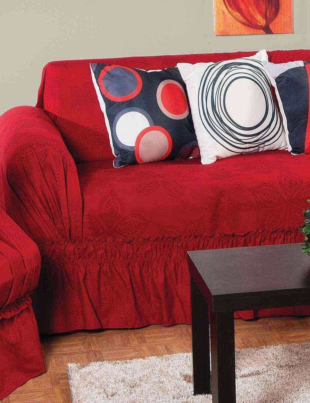A red sofa featuring the Protector de Sala Amapola Sofá by Intima Hogar is adorned with three modern red, black, and white throw pillows. In front, a dark wood coffee table and beige rug sit below a bold red abstract painting.