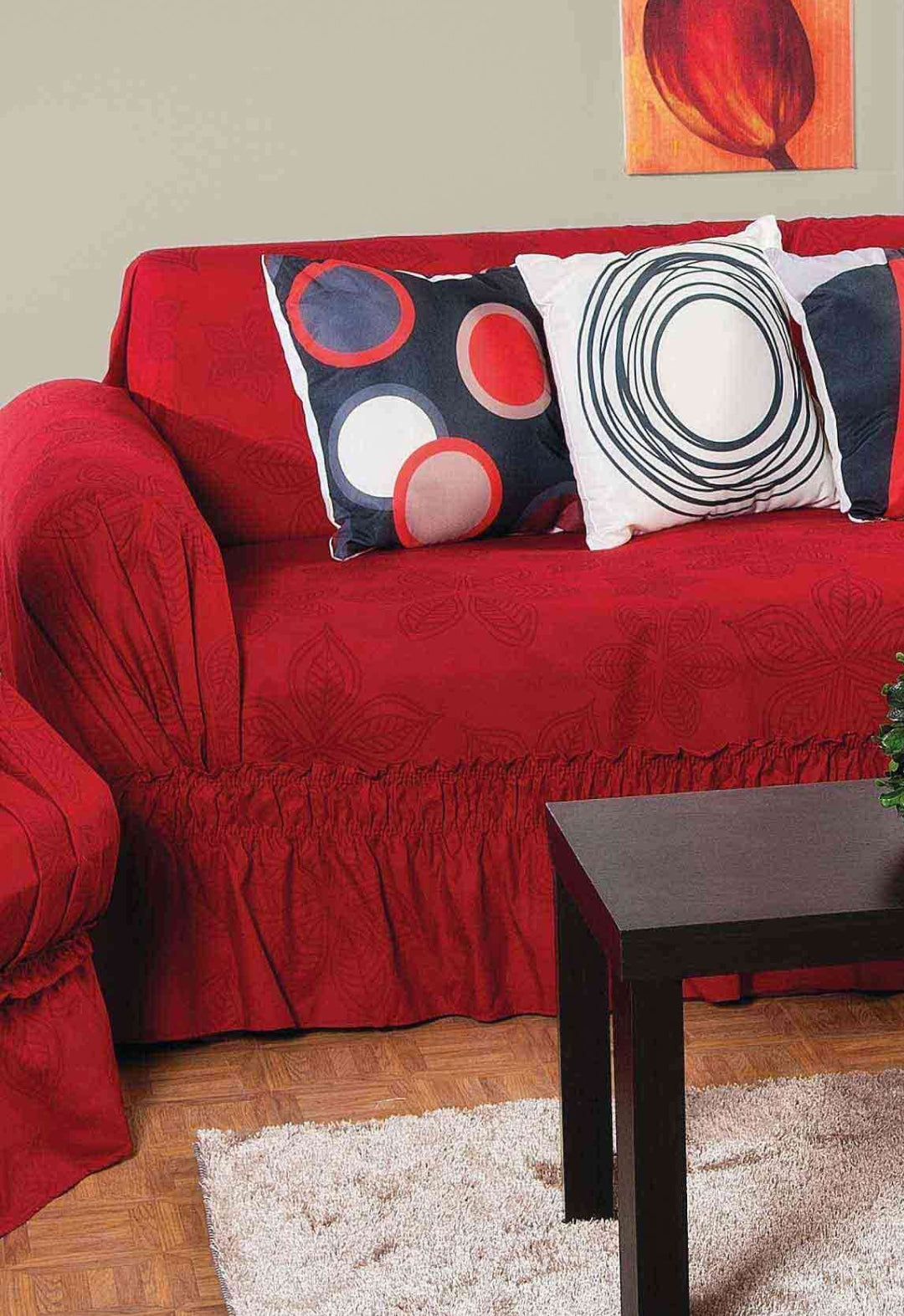 A red sofa featuring the Protector de Sala Amapola Sofá by Intima Hogar is adorned with three modern red, black, and white throw pillows. In front, a dark wood coffee table and beige rug sit below a bold red abstract painting.