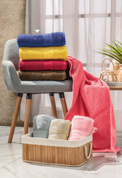 A stack of Juego de 2 Toallas Americana Café by Intima Hogar rests on a gray chair, one towel draped over the side. In front, a basket holds pastel cotton towels as sunlight streams through the window.