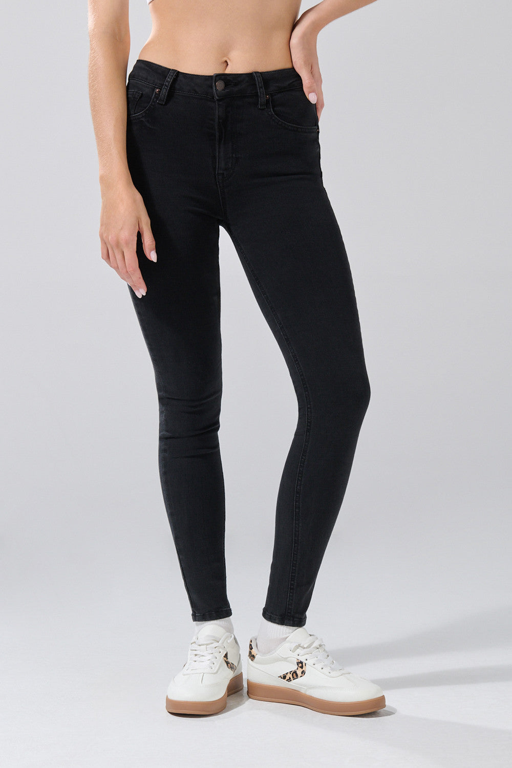 A woman wears KOAJ PANTALON KOAJ JEAN PUSH UP 32008 1/26, high-waisted black skinny jeans by KOAJ, paired with white sneakers featuring leopard print details. The photo is cropped from the waist down against a light gray background.