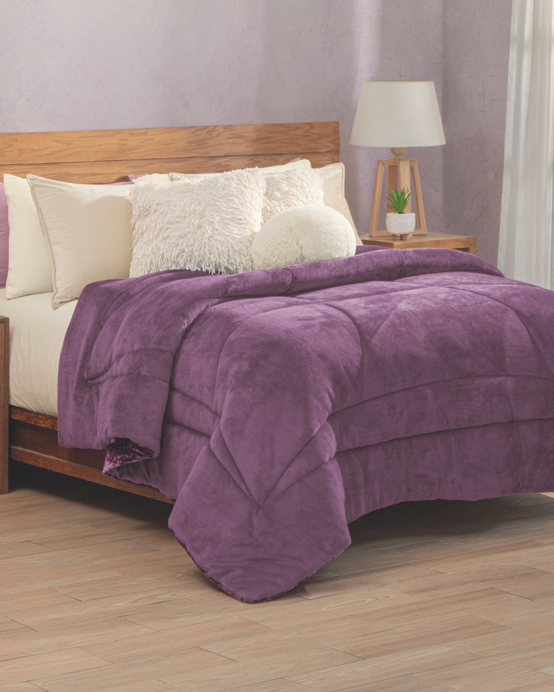 A neatly made bed with cream pillows and a fluffy white cushion, covered by the Cobertor Velvet Ice Deluxe Amatista from Intima Hogar, sits beside a wooden nightstand with a lamp and small plant.