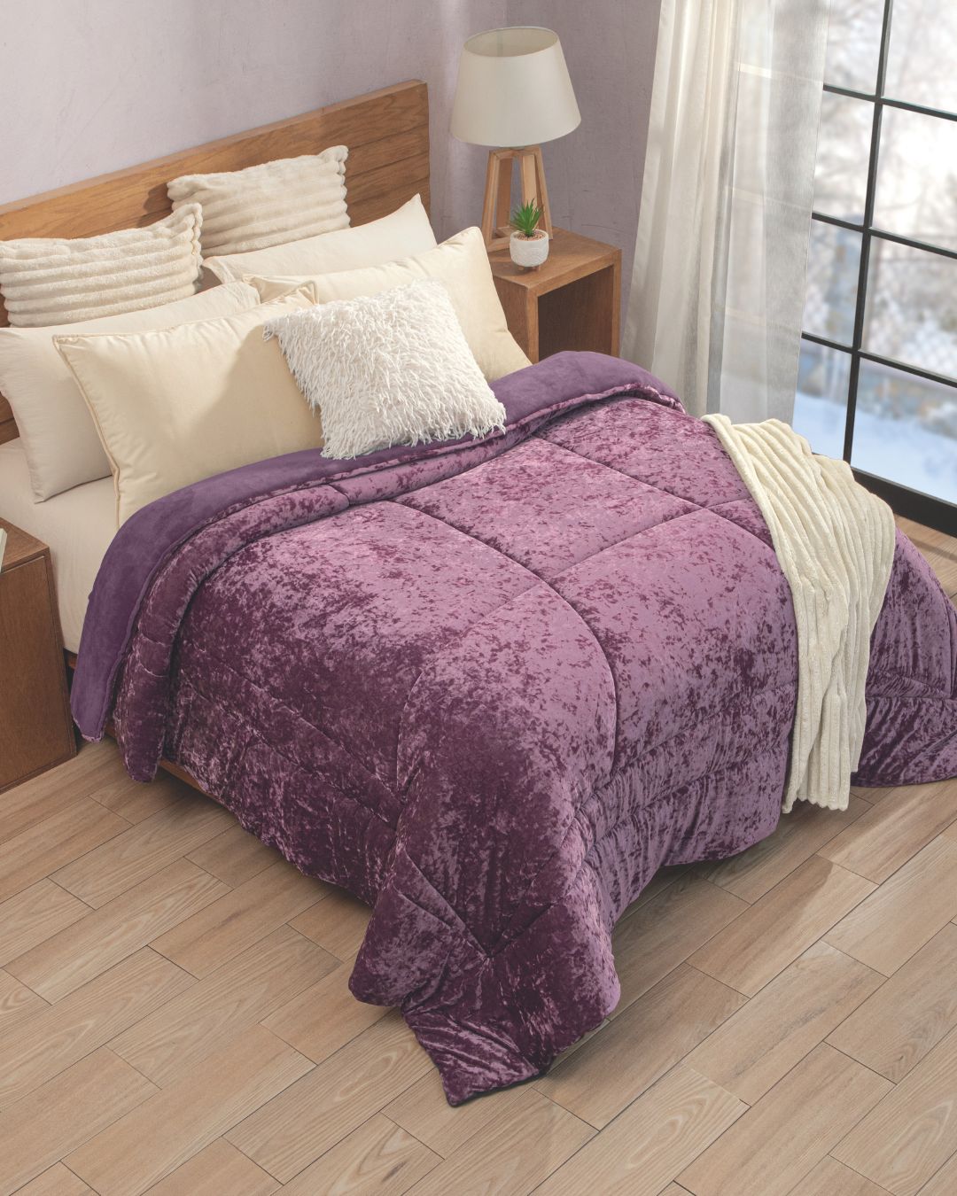 A neatly made bed with cream and white pillows is topped with the Cobertor Velvet Ice Deluxe Amatista from Intima Hogar—a plush purple comforter—and a cream throw, all set in a cozy, sunlit bedroom with wood floors and a lamp on the side table.