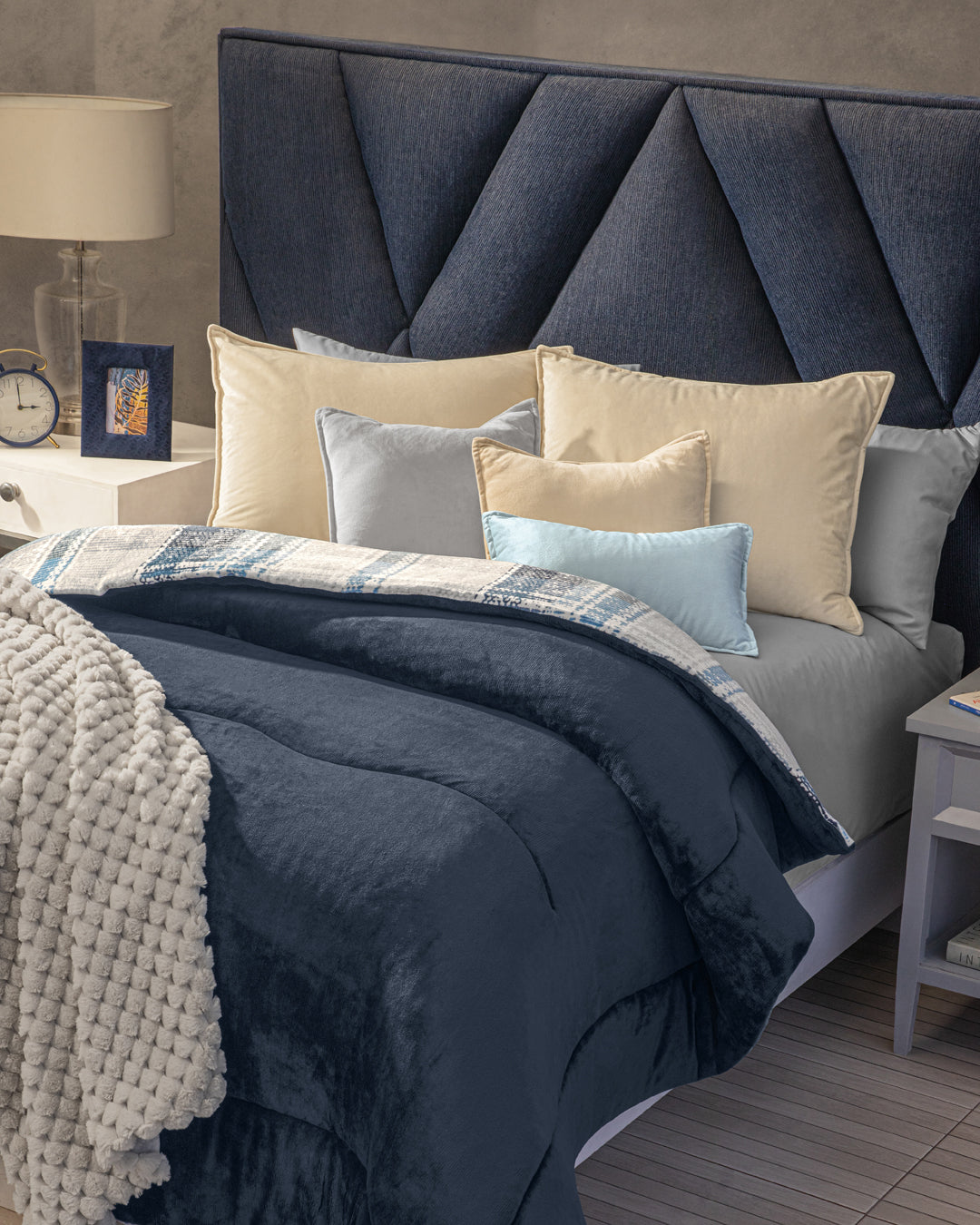 A neatly made bed features the Cobertor Flannel Extra Suave Duque by Intima Hogar in navy blue, complemented by cream and light blue pillows, a knit blanket, and a geometric navy headboard. A nightstand with a lamp, clock, and framed photo sits beside the bed, enhancing the room’s cozy yet sophisticated ambiance.