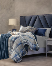 A cozy bedroom featuring the Intima Hogar Cobertor Flannel Extra Suave Duque as a warm blue throw blanket, a modern navy blue headboard, reversible blue and gray plaid bedding, assorted blue and gray pillows, and a nightstand topped with a lamp, books, and clock.