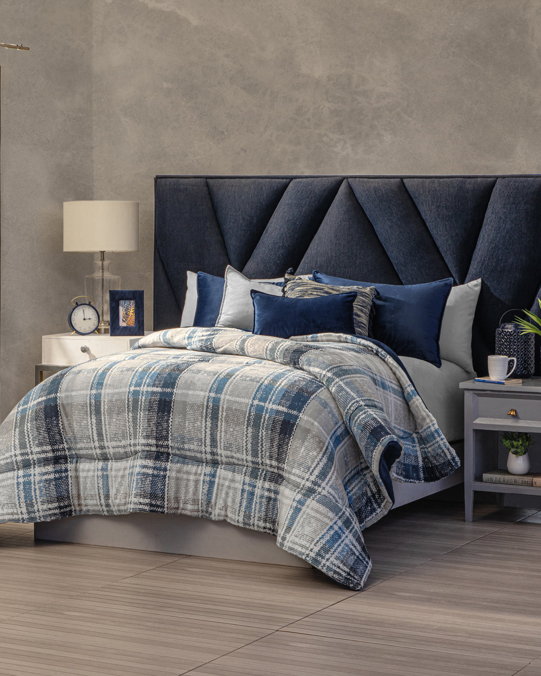 A modern bedroom with a dark blue upholstered headboard, plaid blue and gray bedding featuring the Cobertor Flannel Extra Suave Duque by Intima Hogar, several pillows, two nightstands with lamps, a clock, a mug, and a book, all set against a gray textured wall.