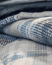 A close-up of the Intima Hogar Cobertor Flannel Extra Suave Duque, showcasing folded, patterned fabric in blue, gray, and white tones with a soft, textured surface and abstract designs. This modelo doble vista offers versatile style and cobertura cálida that feels suave al contacto.