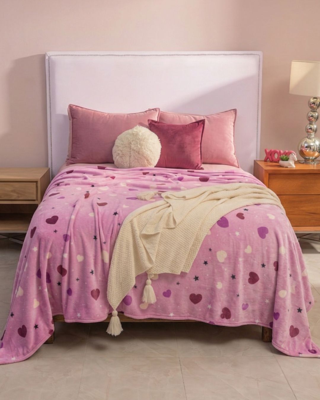 A neatly made bed features the Intima Hogar Cobertor Flannel Ligero Lilian with a pink and purple heart pattern, three pink pillows, a round white cushion, and a cream throw. Wooden nightstands with lamps add charm for cozy nights.