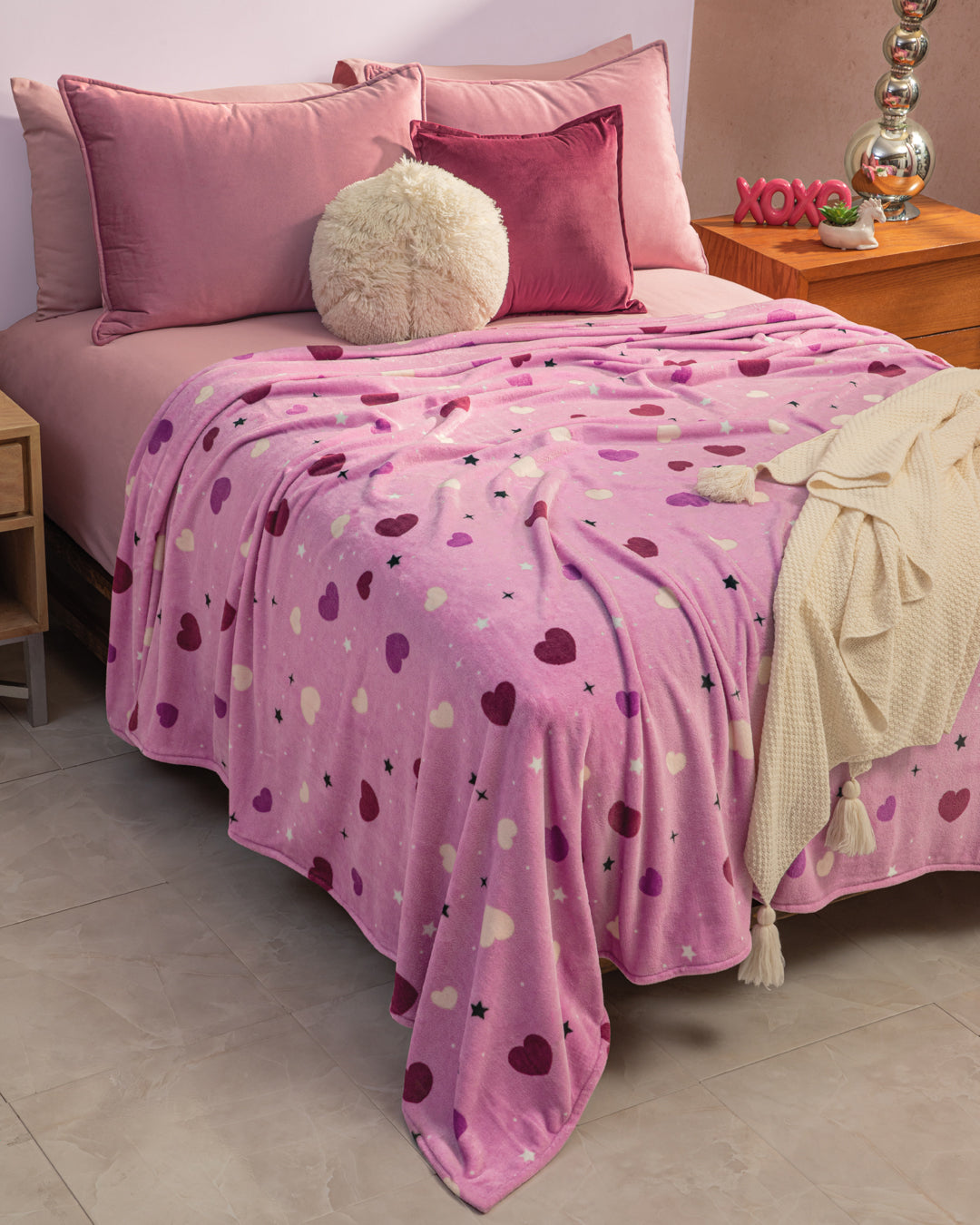 A neatly made bed features the Intima Hogar Cobertor Flannel Ligero Lilian in pink and purple, with matching pillows, a round fluffy cushion, and a cream throw on one side. Nightstands display decor and a lamp.
