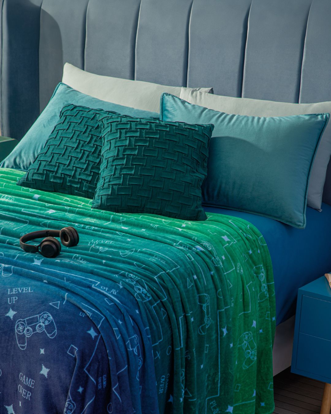 The Cobertor Flannel Ligero Videojuegos de Intima Hogar luce sus estampados gamer, controles y Level Up sobre una cama con almohadas de terciopelo teal y cabecera gris, creando un ambiente ideal para descansar. Audífonos negros completan el look.