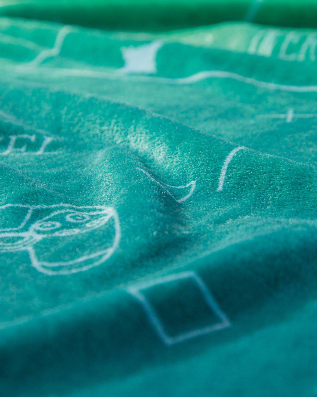 Close-up of the Cobertor Flannel Ligero Videojuegos by Intima Hogar, featuring teal fabric with white line drawings of geometric shapes and a cartoon face, shown in soft lighting to highlight its texture.