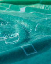 Close-up of the Cobertor Flannel Ligero Videojuegos by Intima Hogar, featuring teal fabric with white line drawings of geometric shapes and a cartoon face, shown in soft lighting to highlight its texture.