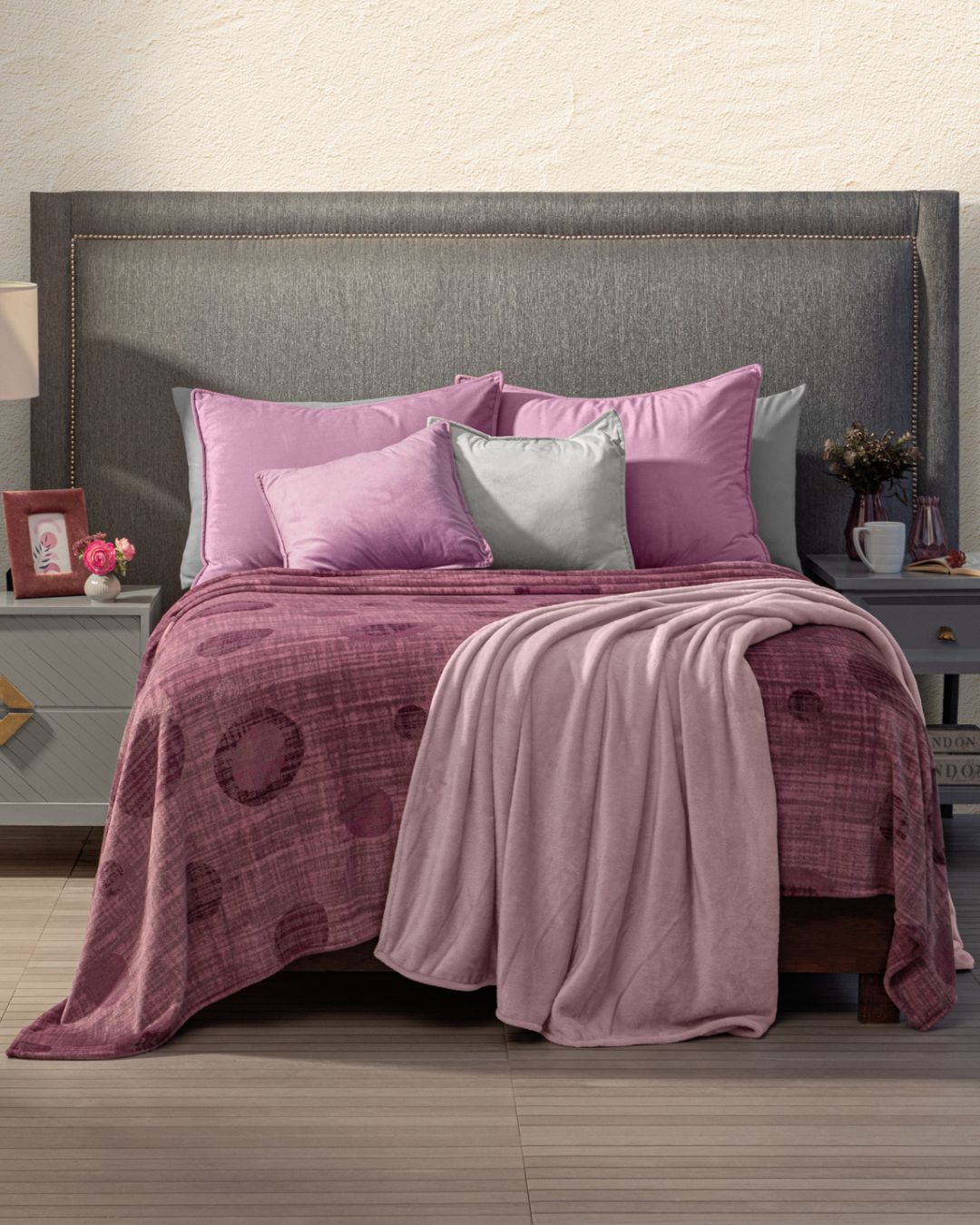 A neatly made bed with a gray headboard, pink and purple pillows, a pink blanket, and the Cobertor Flannel Ligero Marruecos by Intima Hogar highlights diseño cálido. Each side features nightstands with a lamp, frame, clock, and vase.