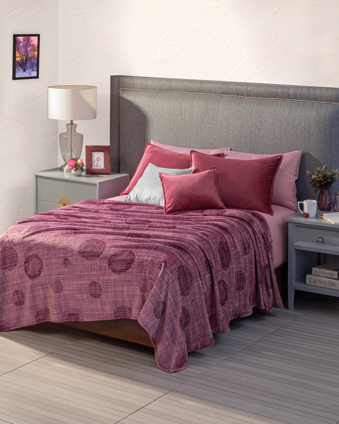 A neatly made bed features the Cobertor Flannel Ligero Marruecos by Intima Hogar, plus matching pillows and a gray upholstered headboard. Each nightstand has a lamp, photo, mug, book, small decor, and a framed picture decorates the wall.