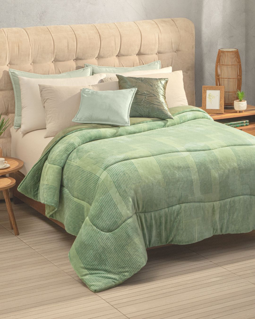 A neatly made bed featuring the Cobertor Flannel Extra Suave Kioto by Intima Hogar, accented with green and beige pillows, a beige tufted headboard, and side tables adorned with plants and decor in a softly lit bedroom, creates a serene and elegant retreat.