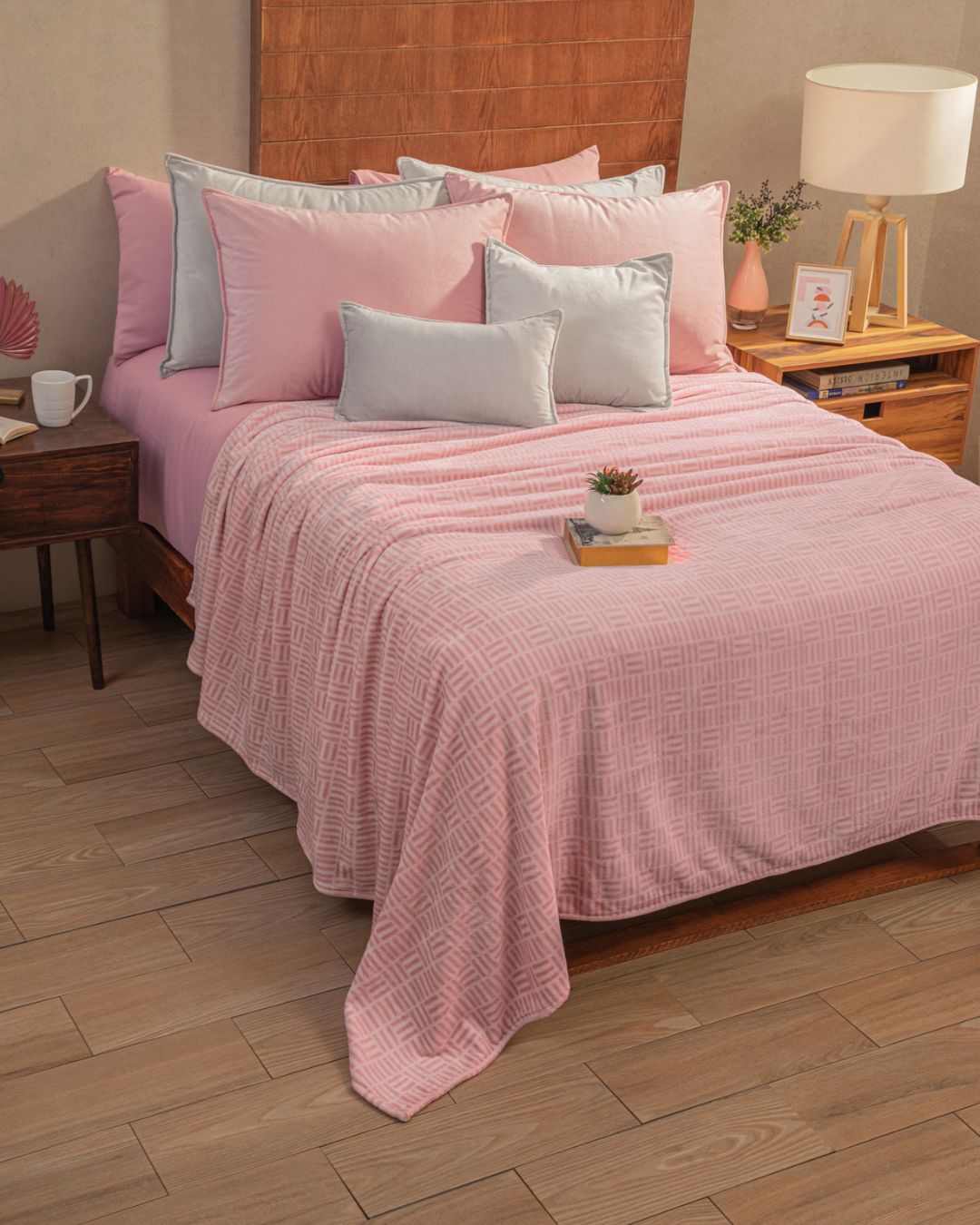 A neatly made bed with the Cobertor Flannel Ligero Jordania by Intima Hogar, featuring a pink quilt and pink-gray pillows rests against a wooden headboard. Each pieza cómoda holds lamps, books, and a small plant over the light wood floor.