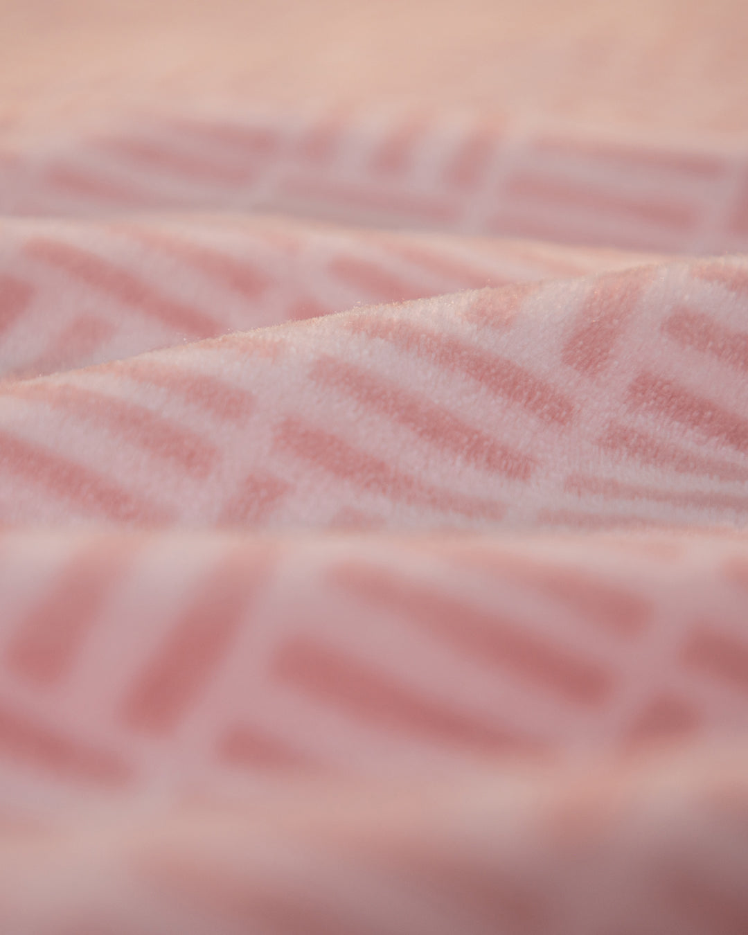 Close-up of Cobertor Flannel Ligero Jordania by Intima Hogar: its soft pink fabric features a geometric pattern with gentle folds and a slightly fuzzy texture, giving a cozy and warm appearance.