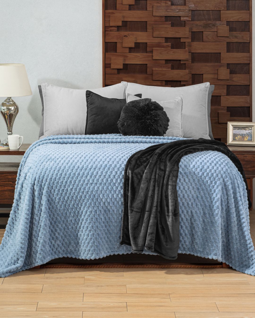 A neatly made cama with the Cobertor Flannel Ligero Niagara de Intima Hogar adds frescura y estilo relajado. Gray and black pillows, a folded black throw, a geometric wooden headboard, and bedside tables with decor complete the look.