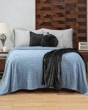 A neatly made cama with the Cobertor Flannel Ligero Niagara de Intima Hogar adds frescura y estilo relajado. Gray and black pillows, a folded black throw, a geometric wooden headboard, and bedside tables with decor complete the look.