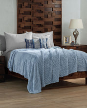 A cozy bedroom with a wooden bed frame, Intima Hogar’s Cobertor Flannel Ligero Niagara in light blue, white and blue accent pillows, a wooden nightstand with lamp, a framed photo, and a mug on a light wood floor.
