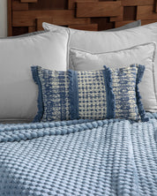 A bed with white pillows, a blue and cream fringed cushion, and the Cobertor Flannel Ligero Niagara by Intima Hogar as a textured light blue blanket, all set against a wooden headboard.