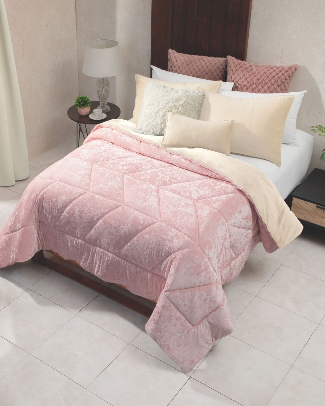 A neatly made bed featuring the Cobertor Velvet Ice Deluxe Loto by Intima Hogar, paired with cream and blush pillows. Next to the bed, a nightstand holds a lamp, a book, and a small plant. The room is finished with tiled floors and soft, neutral-colored walls.
