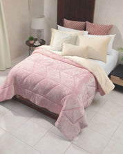 A neatly made bed featuring the Cobertor Velvet Ice Deluxe Loto by Intima Hogar, paired with cream and blush pillows. Next to the bed, a nightstand holds a lamp, a book, and a small plant. The room is finished with tiled floors and soft, neutral-colored walls.