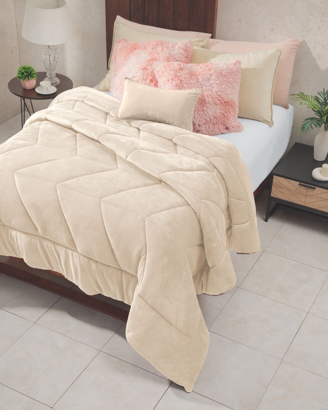 A neatly made bed with the Cobertor Velvet Ice Deluxe Loto by Intima Hogar in cream, accented by pink and beige pillows, a wooden headboard, and bedside tables—one adorned with a lamp and decor, the other with a plant. The room features light tiled flooring.
