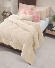 A neatly made bed with the Cobertor Velvet Ice Deluxe Loto by Intima Hogar in cream, accented by pink and beige pillows, a wooden headboard, and bedside tables—one adorned with a lamp and decor, the other with a plant. The room features light tiled flooring.
