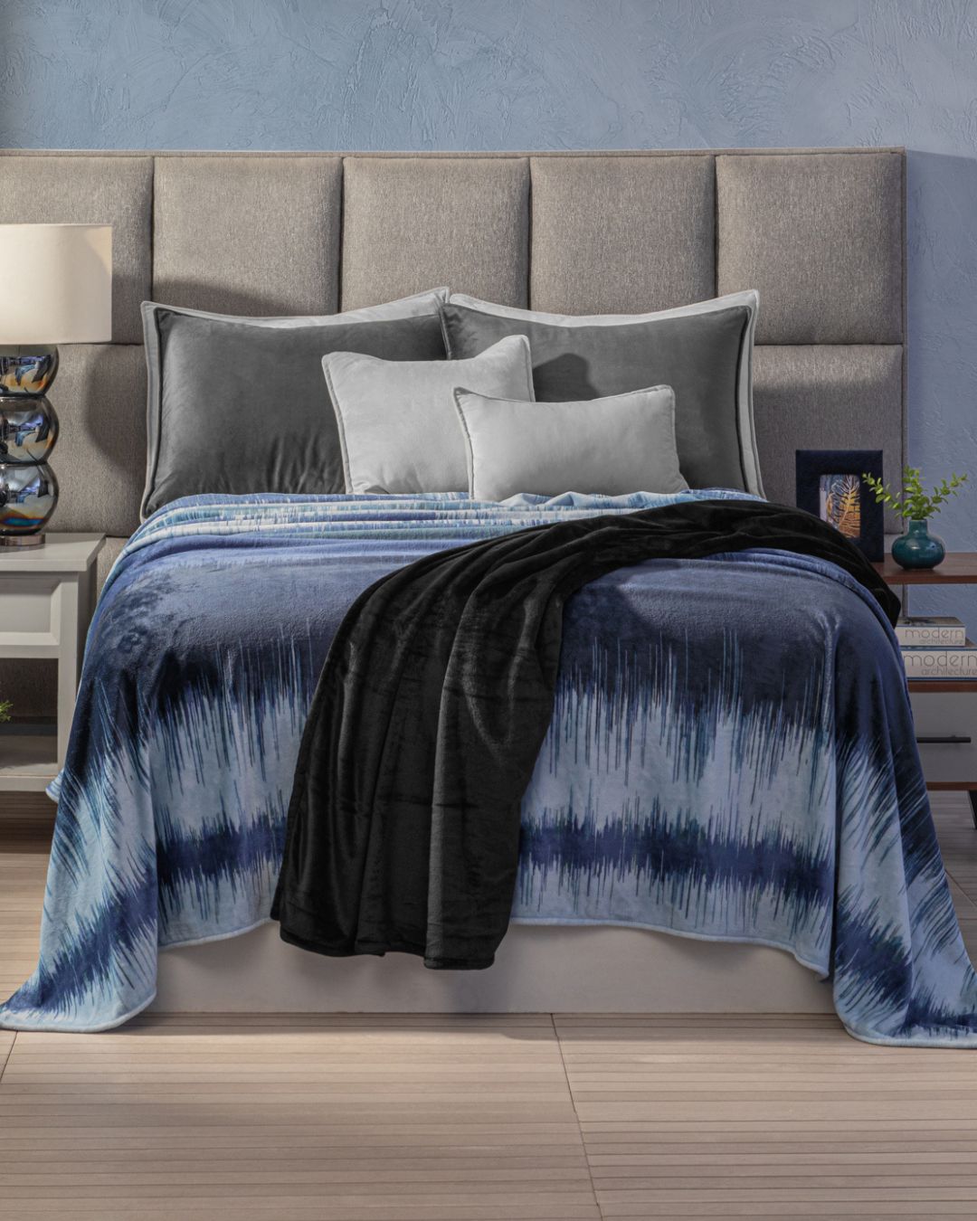 A neatly made bed with a gray upholstered headboard, gray and silver pillows, the Cobertor Flannel Ligero Miami by Intima Hogar, and a black throw creates un diseño fresco. Nightstands with lamps and decor are on each side.