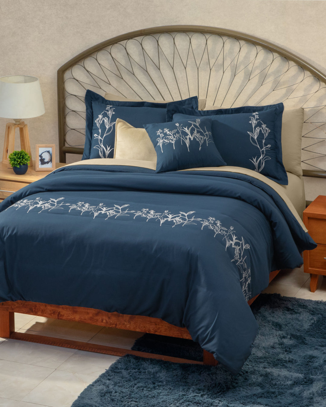 A neatly made bed dressed in the Juego de Duvet Luxury Monarca by Intima Hogar, showcasing navy blue bedding with white floral embroidery, complemented by beige accent pillows and elegant fundas decorativas, a wooden headboard, bedside table, lamp, small plant, and a blue rug on a tiled floor.
