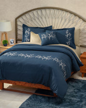 A neatly made bed dressed in the Juego de Duvet Luxury Monarca by Intima Hogar, showcasing navy blue bedding with white floral embroidery, complemented by beige accent pillows and elegant fundas decorativas, a wooden headboard, bedside table, lamp, small plant, and a blue rug on a tiled floor.