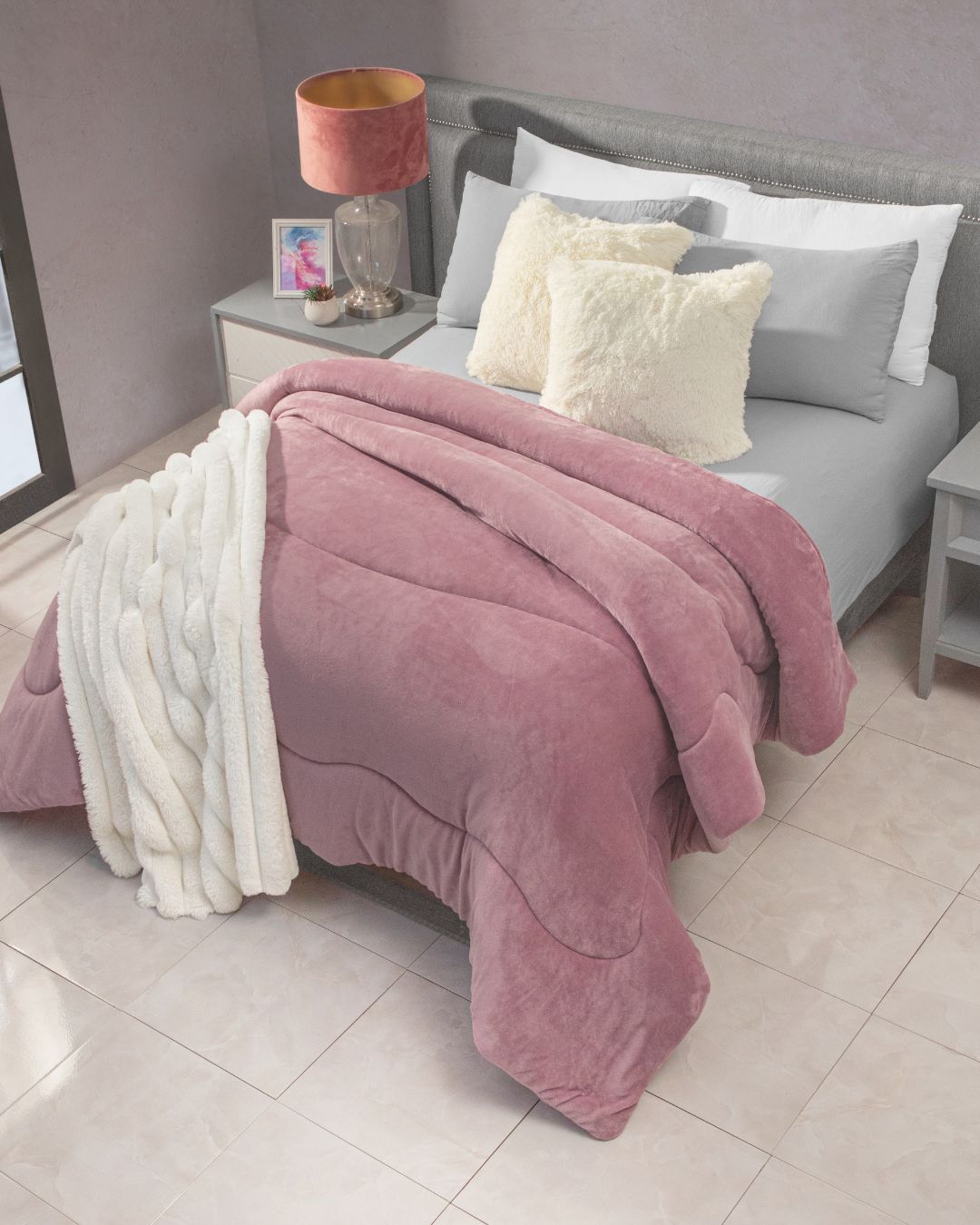 A neatly arranged bed adorned with the Cobertor Flannel Extra Suave Mónaco by Intima Hogar, featuring subtle patterns in soft pink hues, a white blanket, and fluffy pillows creates an inviting dormitorio. Gray side tables with lamps, a photo frame, and glass decor complement the light-colored tiled floors and neutral walls, enhancing la sensación de suavidad throughout the space.