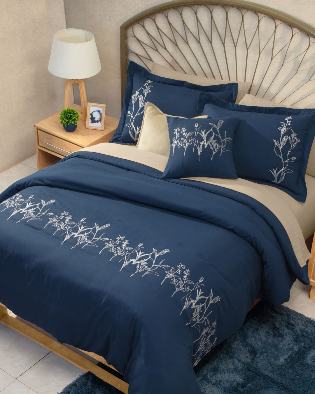 A neatly made bed is dressed with the Juego de Duvet Luxury Monarca by Intima Hogar, showcasing navy blue bedding adorned with elegant white floral patterns and matching fundas decorativas. The look is complemented by an edredón blanco, a wooden headboard, and a nightstand featuring a lamp, potted plant, and framed photo set beside the bed on a blue rug.