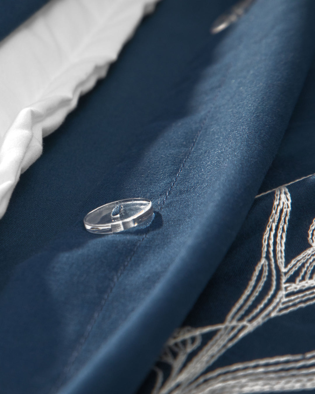 Close-up of the Juego de Duvet Luxury Monarca by Intima Hogar in blue fabric, featuring delicate white embroidery, a clear button, and a glimpse of the white inner lining along the edge—perfect for adding a touch of elegance to your edredón blanco or coordinating seamlessly with your favorite fundas decorativas.