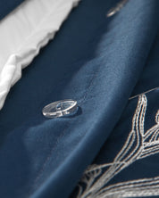 Close-up of the Juego de Duvet Luxury Monarca by Intima Hogar in blue fabric, featuring delicate white embroidery, a clear button, and a glimpse of the white inner lining along the edge—perfect for adding a touch of elegance to your edredón blanco or coordinating seamlessly with your favorite fundas decorativas.